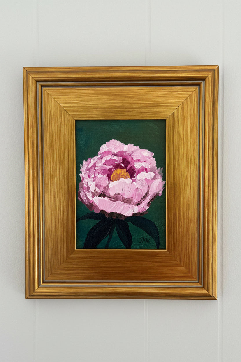 A framed painting of a pink peony flower with dark green leaves on a dark background, enclosed in a wide gold frame and mounted on a white paneled wall.