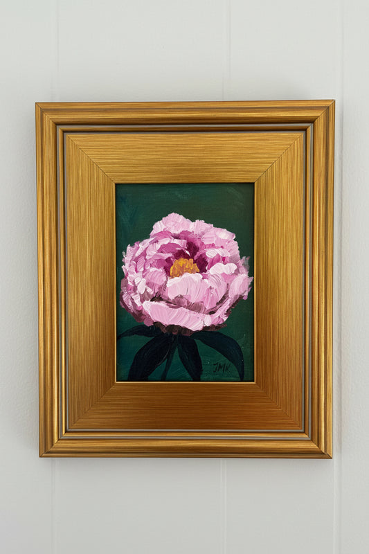A framed painting of a pink peony flower with dark green leaves on a dark background, enclosed in a wide gold frame and mounted on a white paneled wall.